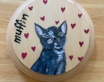 May include: A round wooden plaque with a black and white cat painting. The cat is looking at the viewer with a curious expression. The plaque is decorated with red hearts and the word "Muffin" is written on the left side.
