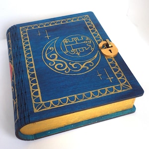 Op de afbeelding: Een blauwe houten doos met een gouden slot en een decoratief ontwerp. De doos is versierd met een halve maan en een symbool in de maan. De doos heeft een gouden rand rond de randen.