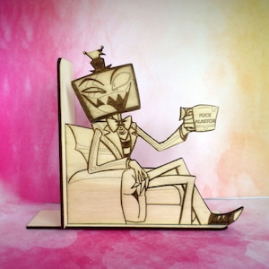 May include: Wooden bookends featuring a cartoon character with a television screen head, seated in a chair and holding a mug. The mug has the text "FUCK ALASTOR". The bookends are made of light-colored wood with dark etched details.