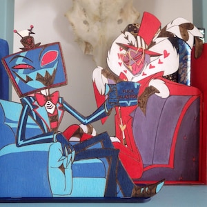 Puede incluir: Dos sujetalibros de madera con personajes de dibujos animados. Un personaje es azul con cabeza en forma de televisor, el otro es rojo y blanco. El personaje azul sostiene un libro con el texto "FUCK ALASTOR". Los sujetalibros están en una estantería.