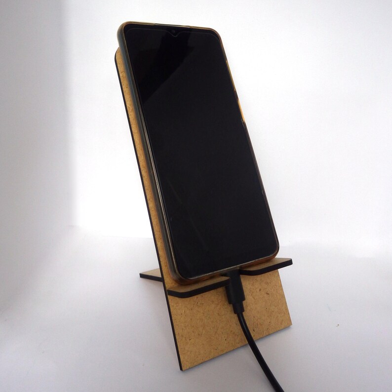 Puede incluir: Un soporte de tel&eacute;fono de madera con un tel&eacute;fono inteligente negro apoyado en &eacute;l. El tel&eacute;fono est&aacute; enchufado con un cable de carga negro.