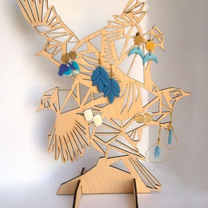 Puede incluir: Soporte para pendientes de madera con forma de bandada de p&aacute;jaros con dise&ntilde;os geom&eacute;tricos. El soporte tiene un acabado marr&oacute;n claro y est&aacute; decorado con pendientes azules, blancos y dorados.