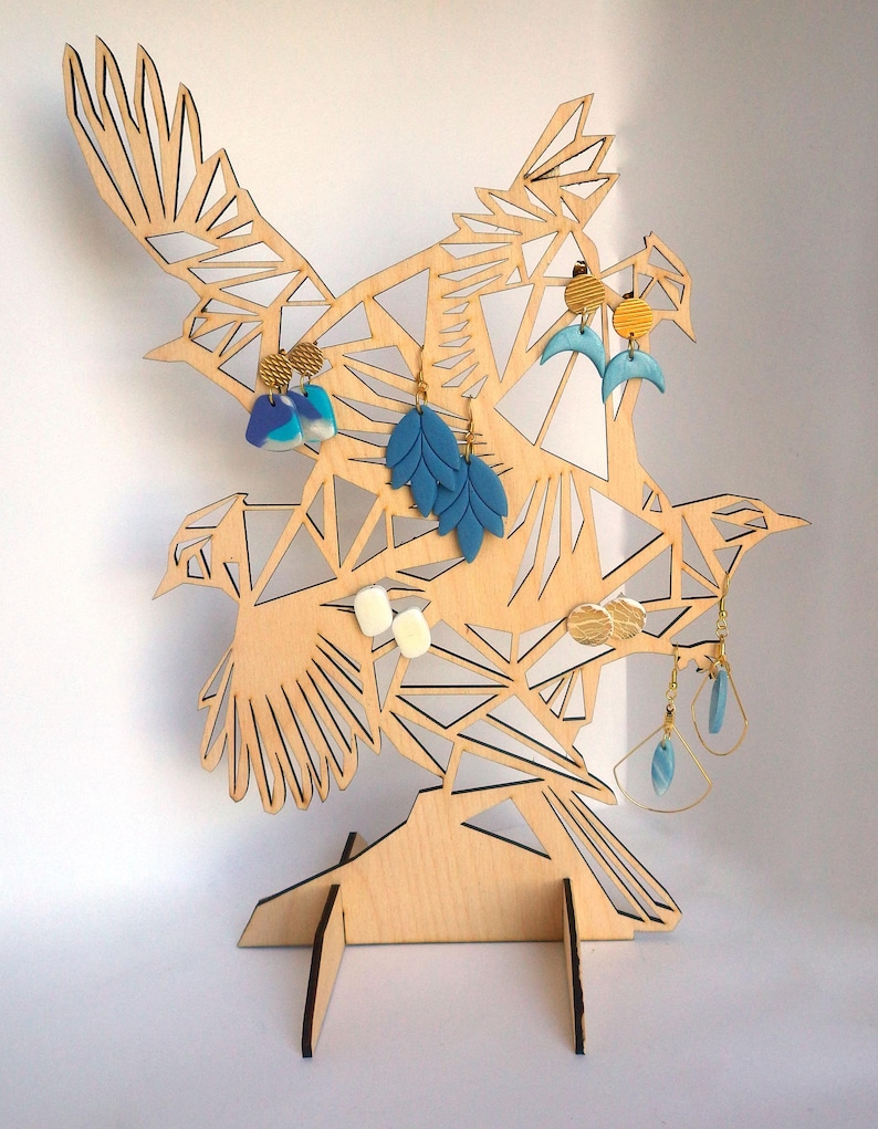 Puede incluir: Soporte para pendientes de madera con forma de bandada de p&aacute;jaros con dise&ntilde;os geom&eacute;tricos. El soporte tiene un acabado marr&oacute;n claro y est&aacute; decorado con pendientes azules, blancos y dorados.