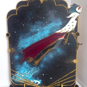 May include: A wooden cutout of a bird with a red cape flying through a starry night sky. The bird is painted in white, red, and black. The background is painted in black with blue and white stars. The cutout is mounted on a wooden base.