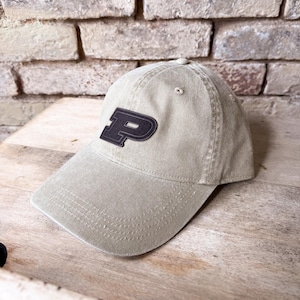 May include: A khaki baseball cap with a black and gray letter "P" patch. The cap has a curved brim and a stitched design. The background is a weathered brick wall and a wooden surface.