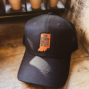 May include: Black trucker hat with a brown leather patch that says "The Corn Fields Are Calling And I Must Go" with an outline of the state of Indiana.