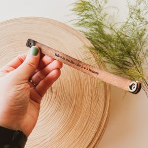 May include: A light brown leather bracelet with the engraved text "Believe in you like I do! 1-4-3 Mommy". The bracelet has metal snap closures. The bracelet is displayed on a wooden surface with green foliage.