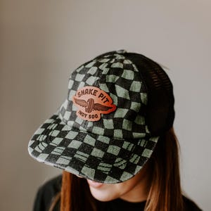 May include: A black and green checkered trucker hat with a mesh back. The hat features a leather patch with the text "Snake Pit Indy 500" and a winged emblem. The hat is worn by a person.