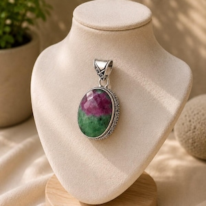 Può includere: Un ciondolo ovale con una pietra di rubino e zoisite sfaccettata, caratterizzata da un mix di colori rosa scuro e verde. La pietra è incastonata in una cornice d'argento decorativa con dettagli in metallo lavorato e un gancio d'argento abbinato.