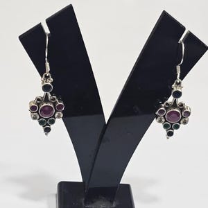 Op de afbeelding: Een paar zilveren bungelende oorbellen met een centrale ronde donkerroze steen omringd door kleinere ronde donkerblauwe, groene en heldere stenen in een clusteropstelling. De oorbellen hangen aan zilveren haken en hebben een metalen afwerking.