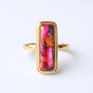 May include: A gold ring featuring a rectangular, vertically oriented inlay of spiny oyster shell. The stone displays a vibrant mix of bright pink, orange, and deep blue hues with metallic gold-colored veining throughout the polished surface.