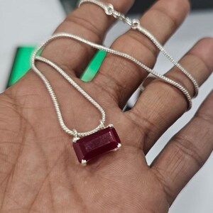 May include: A silver chain necklace featuring a rectangular, deep red gemstone pendant. The pendant is held by a simple silver setting with two prongs on each side, connected to a thin, flexible silver snake chain that loops across the palm.