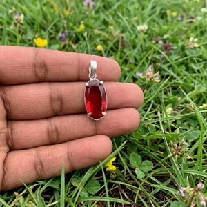 Puede incluir: Un colgante de forma ovalada con una piedra preciosa de color rojo intenso engastada en un marco de metal plateado. El colgante tiene una anilla sencilla en la parte superior para sujetarlo a una cadena, con una superficie lisa y pulida.
