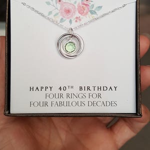 May include: A silver necklace with a pendant of four interlocking rings and a green gemstone. The necklace is in a box with a white card that reads "Happy 40th Birthday Four Rings for Four Fabulous Decades".