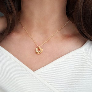 May include: A gold-colored necklace with a pendant featuring interlocking circles. The necklace is worn against a white top, with the chain resting on the chest. The pendant is the focal point, showcasing a delicate design.
