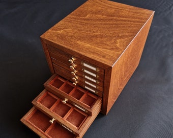 Op de afbeelding: Een houten sieradenkist met meerdere lades, waarvan sommige open zijn. De kist is gebeitst in een warme bruine kleur, met messing knoppen en etikethouders. De binnenkant van de lades is bekleed met roodbruine stof.