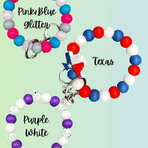 Può includere: Tre portachiavi da polso con perline in rosa, blu e glitter, rosso, bianco e blu, e viola e bianco. I portachiavi hanno una chiusura argentata e un ciondolo a forma di stato del Texas.