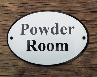 May include: Oval white sign with black border and the words "Powder Room" in black lettering. The sign has two small holes for mounting and is set against a wooden background.