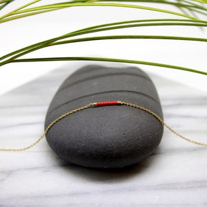 Puede incluir: Un delicado collar de cadena dorada con un pequeño acento de cuentas rojas. El collar se muestra sobre una piedra gris.