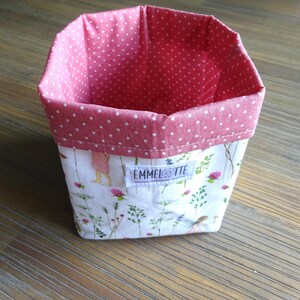 Könnte beinhalten: Ein rosa-weißer Stoffaufbewahrungskorb mit Blumendruck und dem Text "EMMELOTTE" auf der Vorderseite.