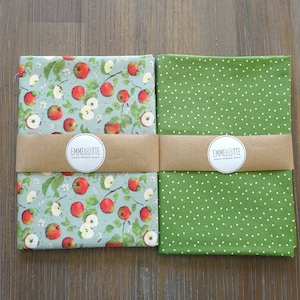 Könnte beinhalten: Zwei Stoffbahnen mit einem braunen Etikett, auf dem "Emmelotte" in weißer Schrift steht. Die Bahn links hat einen hellblauen Hintergrund mit einem sich wiederholenden Muster aus roten Äpfeln und grünen Blättern. Die Bahn rechts hat einen grünen Hintergrund mit einem weißen Punkte-Muster.