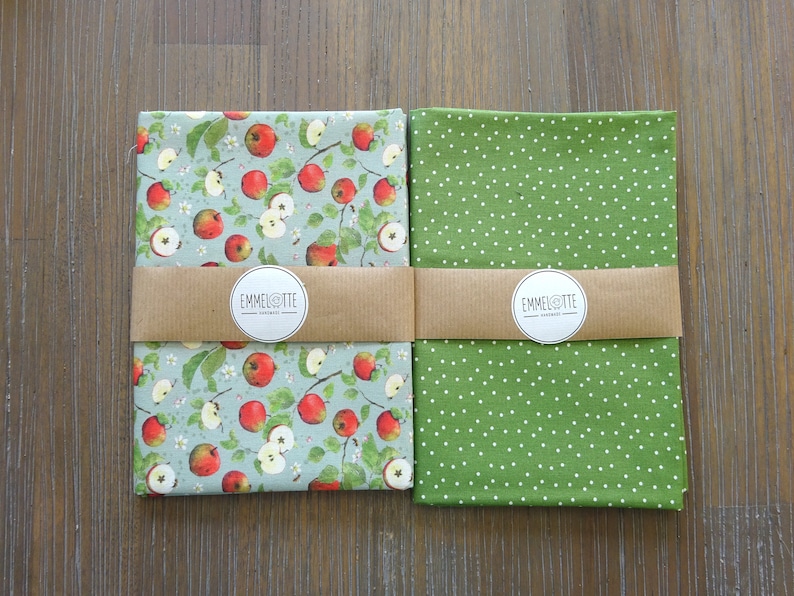 May include: Two fabric panels with a brown label that reads "Emmelotte" in white text. The panel on the left has a light blue background with a repeating pattern of red apples and green leaves. The panel on the right has a green background with a white polka dot pattern.