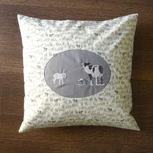 Op de afbeelding: Een wit kussen met een grijze ovale patch in het midden. De patch heeft een geborduurd ontwerp van een moeder schaap en een lam. De kussenstof is bedekt met een herhalend patroon van kleine witte schapen.