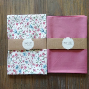 Könnte beinhalten: Zwei gefaltete Stoffservietten. Eine ist weiß mit einem Blumenmuster aus rosa und blauen Blumen. Die andere ist einfarbig rosa. Beide sind mit einem braunen Band mit dem Text "EMMELOTTE" umwickelt.