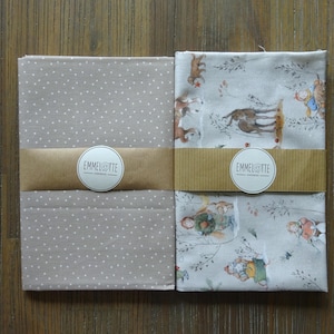 Könnte beinhalten: Zwei Stoffpakete, eines mit einem hellgrauen Polka-Dot-Muster und das andere mit einem skurrilen Waldszenen-Druck. Jedes Paket ist mit einem braunen Papierband und einem weißen Aufkleber mit der Aufschrift "EMMELOTTE" umwickelt.