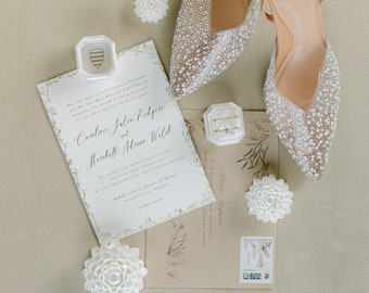 May include: A wedding invitation suite with a white background and gold lettering. The invitation features a floral design and the names "Candice Julie Rodgers" and "Marshall Alan Wild". The invitation is accompanied by a white envelope, a white flower, and a pair of white sparkly heels.