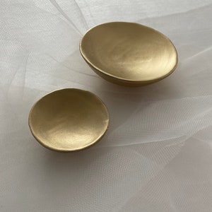May include: Two gold-colored ceramic dishes, one larger than the other, on a white sheer fabric background.