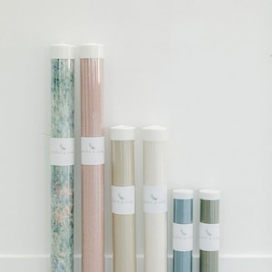 May include: Six rolls of wallpaper in various colors and patterns, including green, pink, beige, white, blue, and green, are displayed in clear plastic tubes. Each tube has a white label with the text "Olive & Oak".