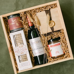 May include: A wooden gift box filled with a bottle of red wine, a jar of apple butter, a box of cookies, and a small tin of peanuts. The box is lined with brown paper shreds.