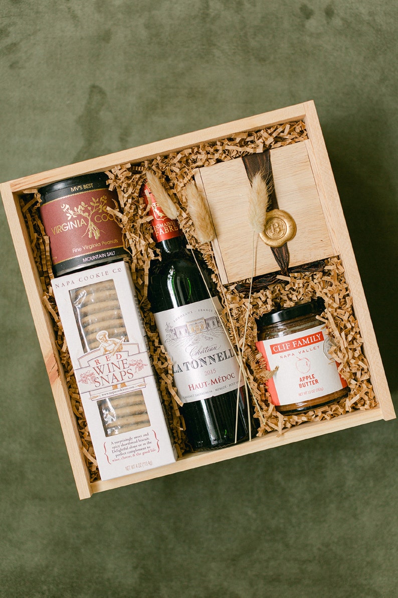 May include: A wooden gift box filled with a bottle of red wine, a jar of apple butter, a box of cookies, and a small tin of peanuts. The box is lined with brown paper shreds.