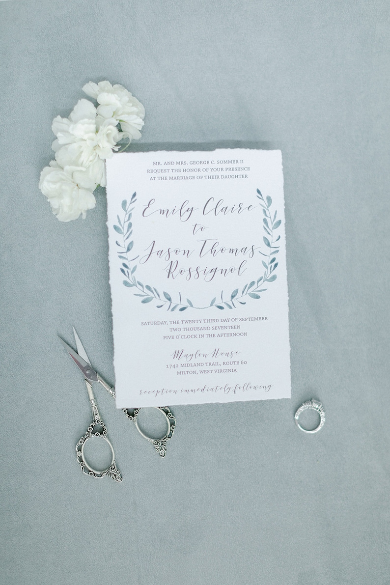 May include: A wedding invitation with a watercolor wreath design in shades of blue. The invitation announces the marriage of Emily Claire to Jason Thomas Rossignol on September 23, 2017, at 5:00 PM at Maglen House, 1742 Midland Trail, Route 60, Milton, West Virginia. The reception will immediately follow.