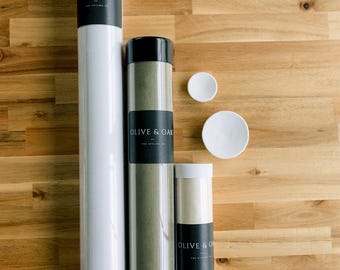 May include: Three cylindrical storage tubes with black caps and black labels reading OLIVE & OAK THE STYLING CO. The tubes are white, olive green, and cream. Two small white circular discs are positioned to the right of the tubes.