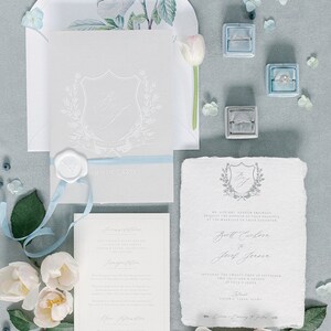 May include: A wedding invitation suite with a gray and white color scheme. The invitation features a crest with the initials "B & J" and the date "MMMDCCXXIV". The invitation is tied with a light blue ribbon. The invitation is surrounded by white flowers and blue berries.