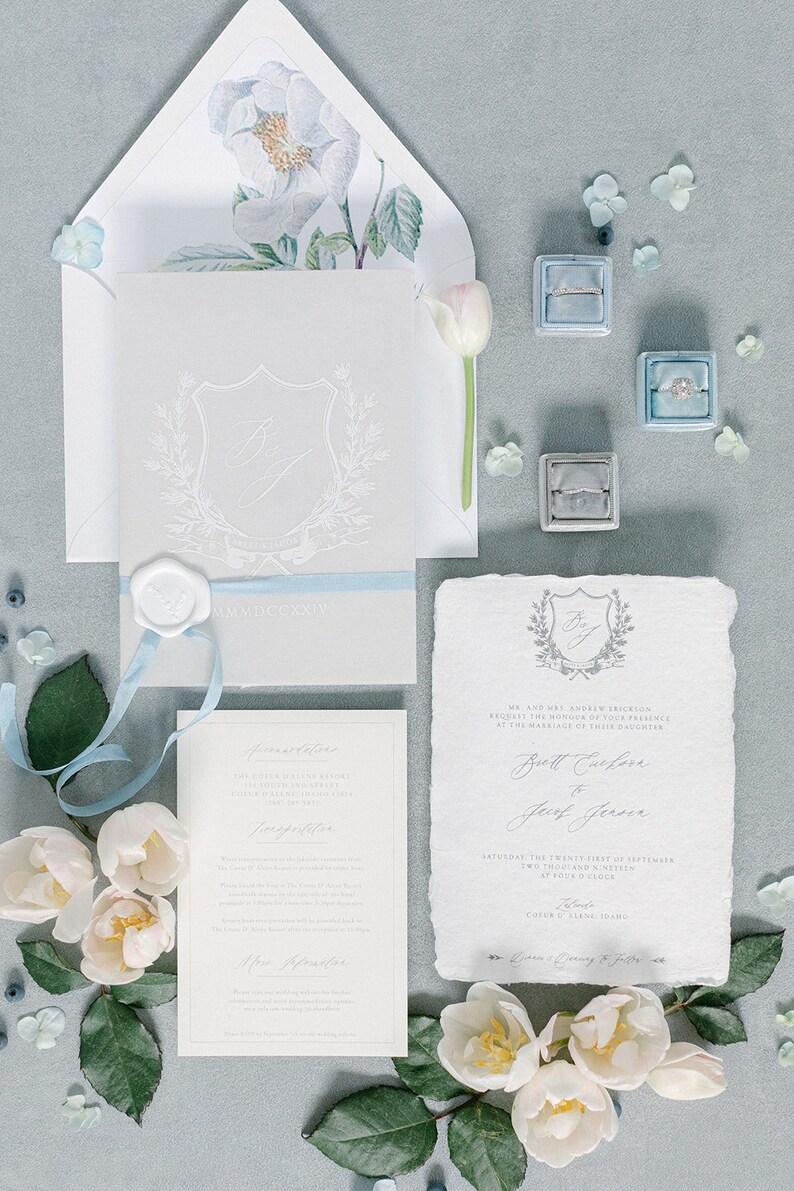 May include: A wedding invitation suite with a gray and white color scheme. The invitation features a crest with the initials "B & J" and the date "MMMDCCXXIV". The invitation is tied with a light blue ribbon. The invitation is surrounded by white flowers and blue berries.