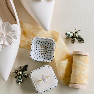 May include: An overhead shot features white bridal shoes with flower accents, a ring box with a gold ring, and bee-shaped brooches. A spool of thread and a ribbon are also present, creating a wedding theme.