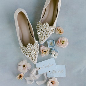 May include: A pair of white satin bridal shoes with pearl embellishments, two small gift boxes, a pair of earrings, and two tags with the names "Caroline" and "Marshall" written in calligraphy.