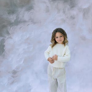 以下が含まれることがあります： クリーム色のリブ編みセーターとストライプのパンツを着た子供。パンツはドローストリングウエストです。背景は、グレーと白の色合いの柔らかい雲模様です。