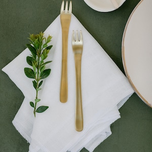 May include: Two gold forks on a white linen napkin with a sprig of green foliage. The napkin is folded in a square shape and is on a green surface.