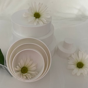 May include: A set of four white round cake stands with a white daisy on top of the largest stand. The stands are stacked on top of each other, with the smallest stand on top. There is a white daisy on the smallest stand.
