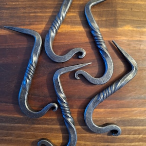 May include: Five hand-forged metal hooks with a twisted handle design. The hooks are dark grey and have a curved shape with a pointed end. The hooks are arranged on a wooden surface.