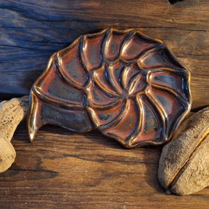 May include: A ceramic dish shaped like a seashell with a spiral design. The dish is glazed in a dark brown and copper color.