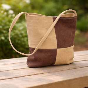 Könnte beinhalten: Eine Patchwork-Tragetasche in Beige- und Brauntönen liegt auf einer Holzbank. Die Tasche hat ein quadratisches Muster und einen langen Riemen. Der Hintergrund ist eine verschwommene Ansicht von grünem Laub und Sonnenlicht.