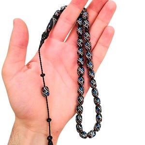 May include: A string of prayer beads featuring dark, oval-shaped beads with a glossy finish. Each bead is decorated with intricate white, red, and blue painted patterns. The beads are connected by a thin black cord, ending in a decorative tassel piece.