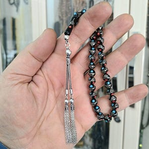May include: A prayer bead strand featuring black spherical beads decorated with small white, red, and light blue dots. The strand includes a silver-toned metal connector and a double silver chain tassel hanging from the end of the bead sequence.