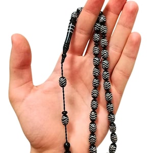 May include: A string of prayer beads featuring black oval-shaped beads with white wavy line patterns and small white dots. The strand includes a long, smooth black cylindrical marker bead with white horizontal lines and a black tassel-like end piece.