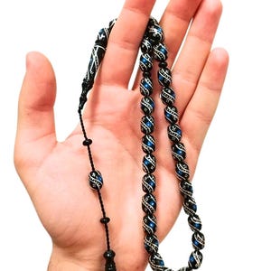 Può includere: Una corona di grani da preghiera composta da perline cilindriche nere con accenti blu e motivi a linee bianche decorative. Le perline sono collegate da un cordino nero e terminano con un pendente nero più lungo e affusolato con dettagli coordinati.